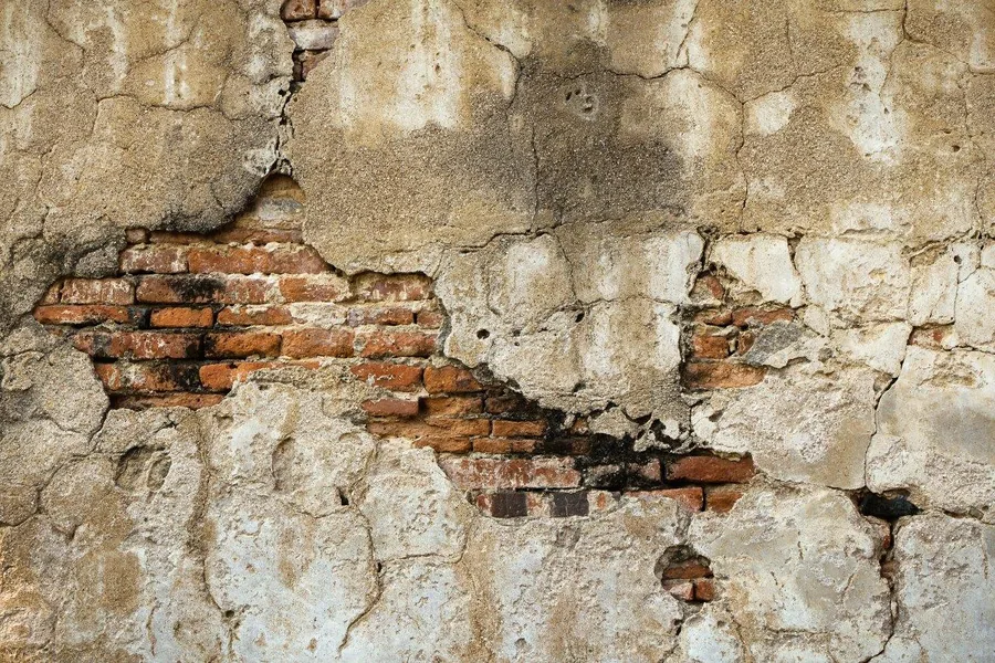 Deteriorating cream plaster revealing aged red brick underneath with natural cracks and wear.