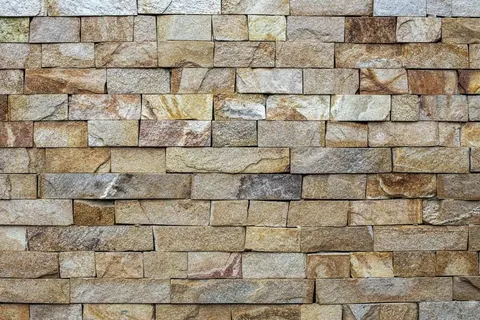 Repeating pattern of stacked rectangular bricks and stones in tan, brown, and grey tones with realistic shadows and texture.
