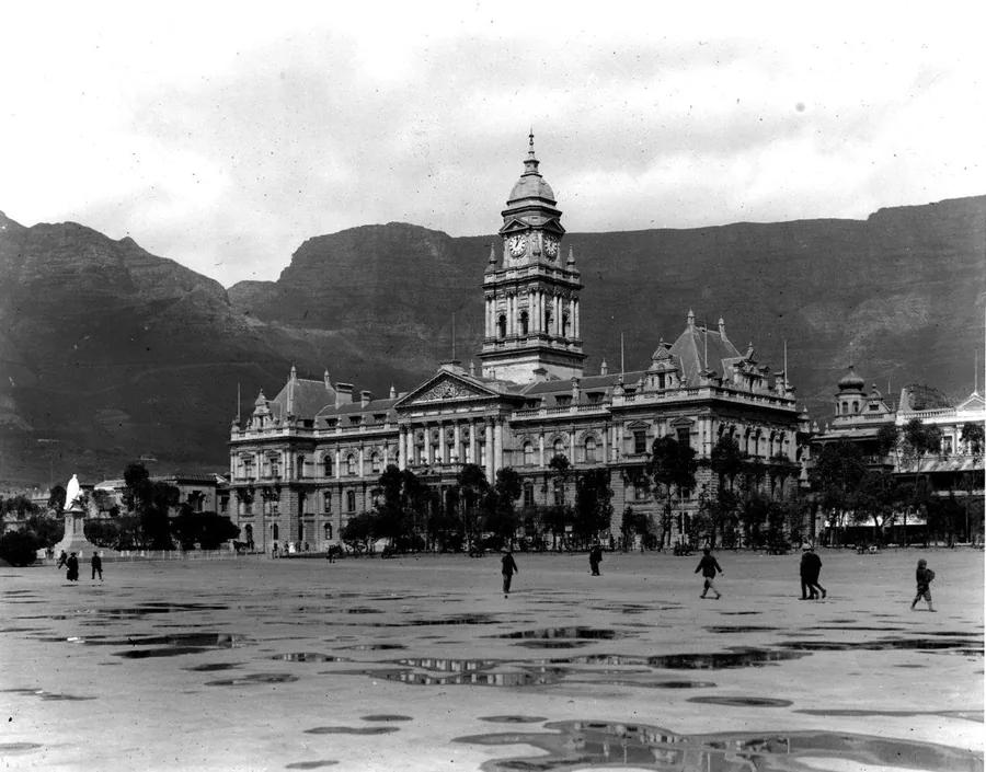 Vintage black and white photograph of Cape Town's grand civic building with clock tower, mountains in background, and wet plaza in foreground.