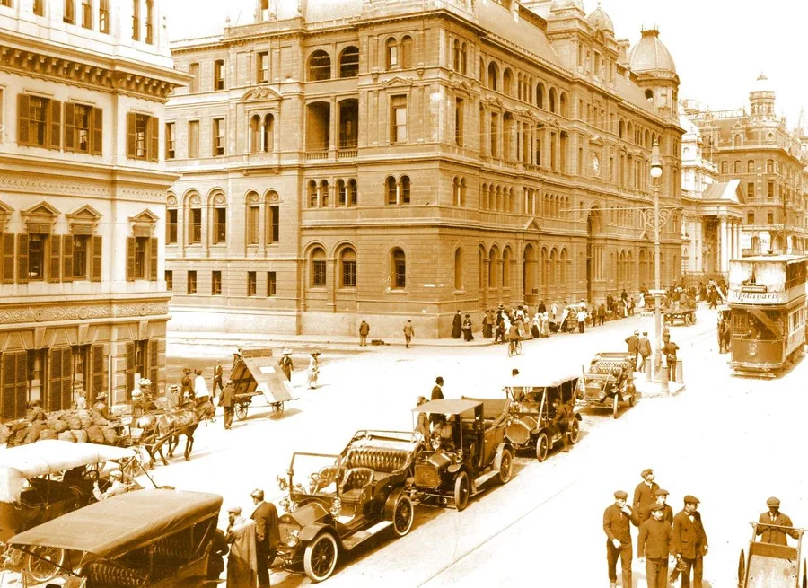 Sepia-toned vintage illustration of colonial-era cityscape with grand architecture, vintage vehicles, and period figures