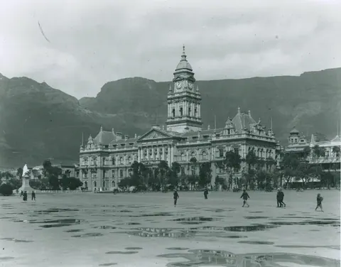 Vintage black and white photograph of Cape Town City Hall with mountains and figures in a plaza.