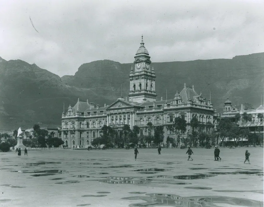 Vintage black and white photograph of Cape Town City Hall with mountains and figures in a plaza.