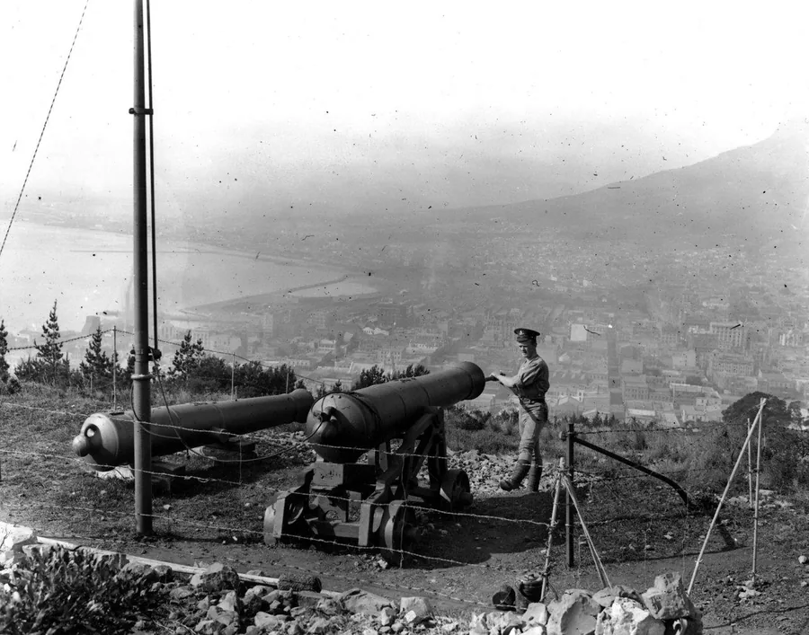Black and white vintage photo of a cannon on a hillside overlooking a dense city valley with mountains in the background.