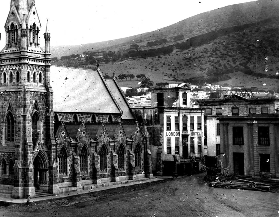 Black and white vintage photograph of Gothic church with colonial buildings and mountain landscape, Cape Town historical street scene.