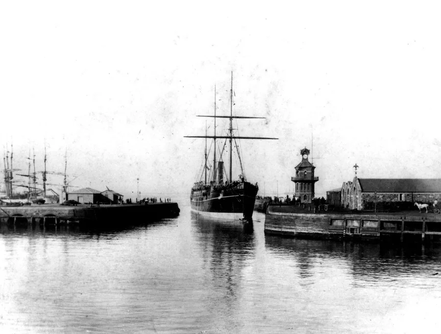 Black and white vintage photograph of steamship in historic harbor with sailing ships and lighthouse.
