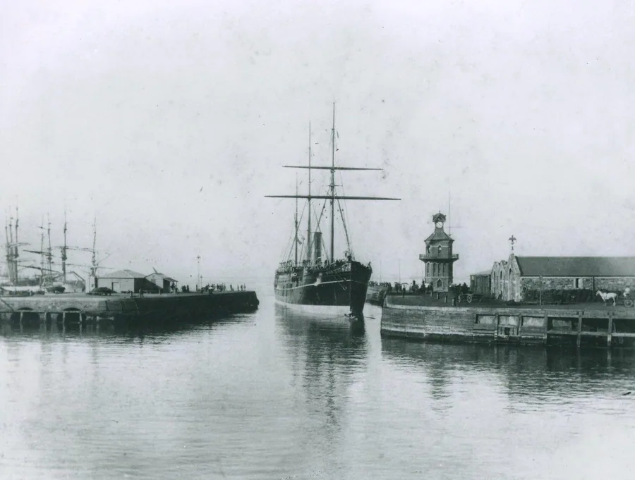 Vintage black and white photograph of a tall ship docked in a historic harbour with lighthouse and colonial buildings.