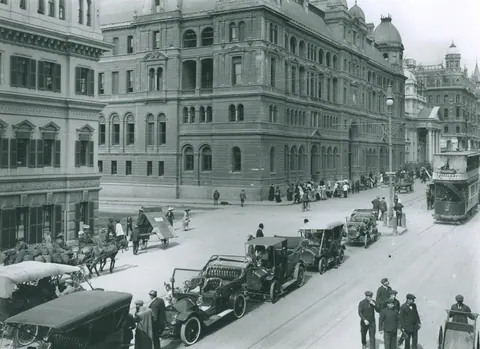 Black and white vintage photograph of a historic Cape Town street with colonial buildings, vintage cars, and pedestrians