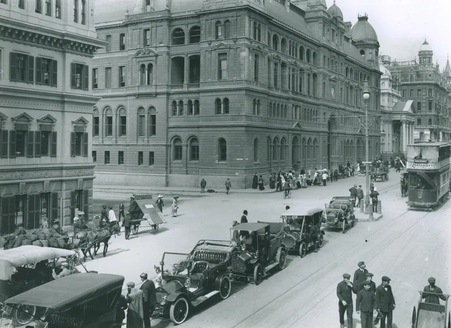 Black and white vintage photograph of a historic Cape Town street with colonial buildings, vintage cars, and pedestrians