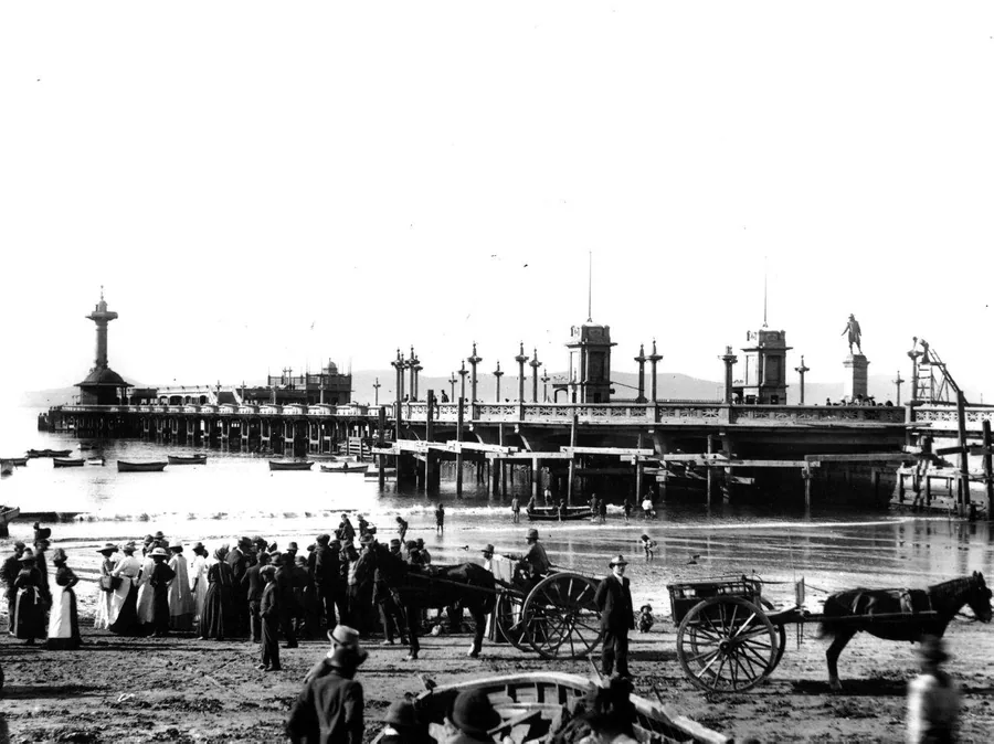 Black and white vintage photograph of a Victorian-era pier with ornamental towers, crowds of people, and horse-drawn carts on the beach.