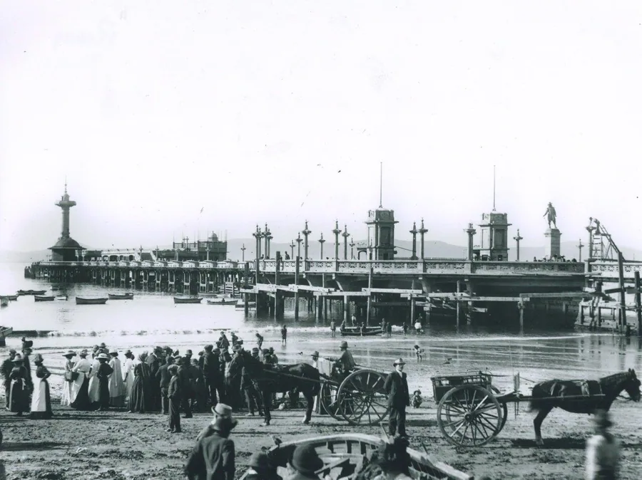 Vintage black and white photograph of a crowded Victorian pier with ornate architecture, horse-drawn carriages, and period crowds.