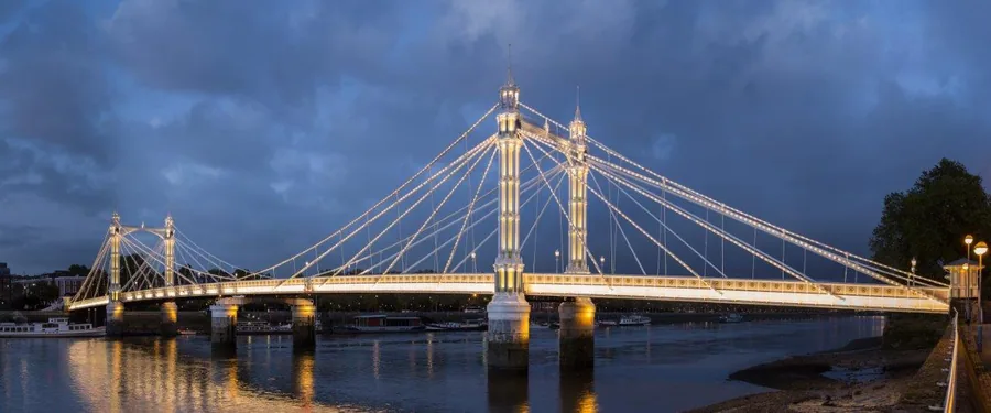 Illuminated suspension bridge at dusk with golden lights reflected in still water under a blue twilight sky.