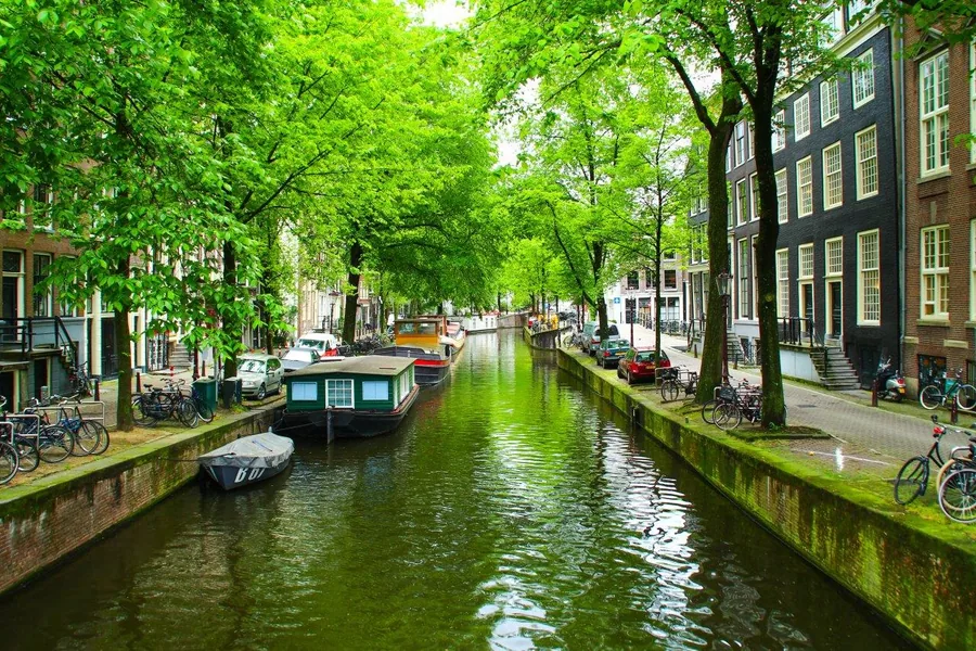 Scenic canal with green trees, historic brick buildings, moored boats, and bicycles along waterfront