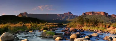 Panoramic view of mountain ranges with blue river, boulders, and golden grass at sunset