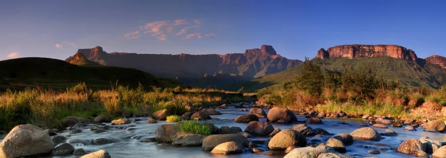 Panoramic view of mountain ranges with blue river, boulders, and golden grass at sunset