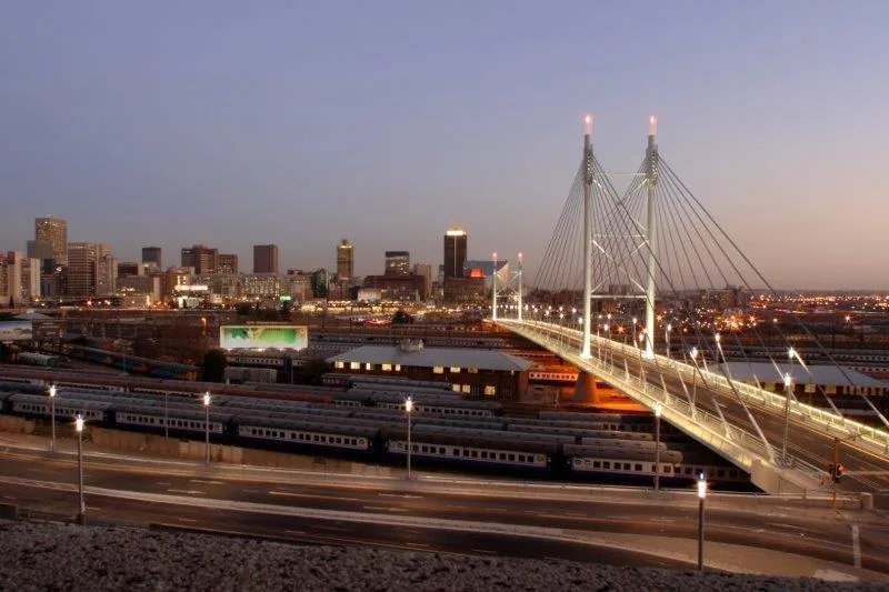 Twilight cityscape with illuminated cable-stayed bridge, trains, and glowing downtown skyline at dusk.