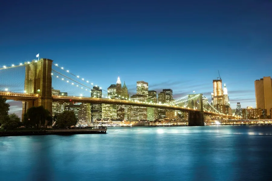 Brooklyn Bridge lit up at dusk with Manhattan skyline reflected in blue water under clear sky