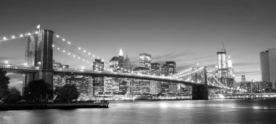 Black and white nighttime cityscape with illuminated Brooklyn Bridge and Manhattan skyline reflected in water.