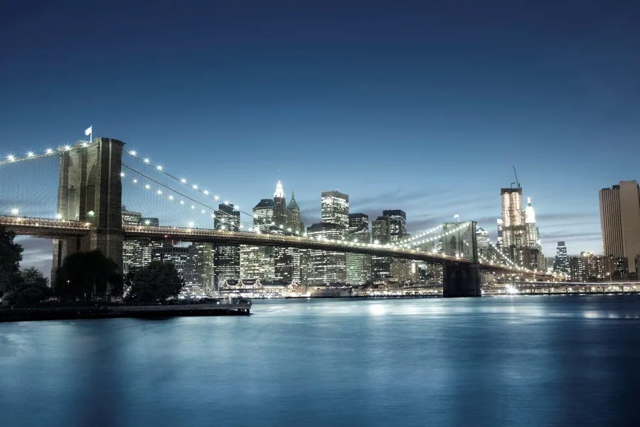 Nighttime view of Brooklyn Bridge and Manhattan skyline with glowing lights reflecting on dark blue water