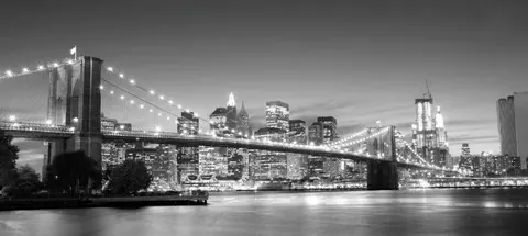 Black and white photograph of Brooklyn Bridge and NYC skyline at night with glowing city lights reflected in water.