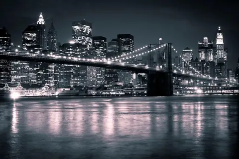 Nighttime New York City skyline with glowing Brooklyn Bridge spanning across dark water, skyscrapers lit against night sky.