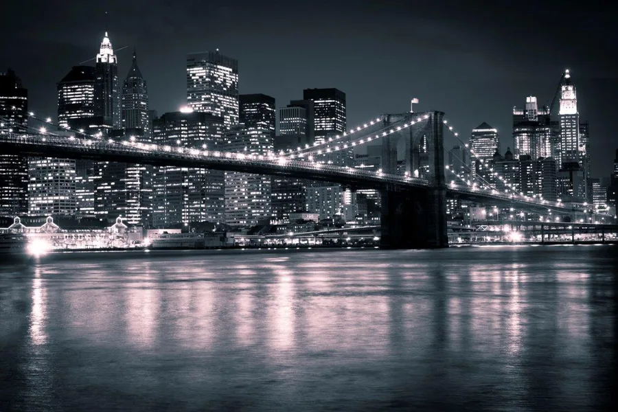 Nighttime New York City skyline with glowing Brooklyn Bridge spanning across dark water, skyscrapers lit against night sky.