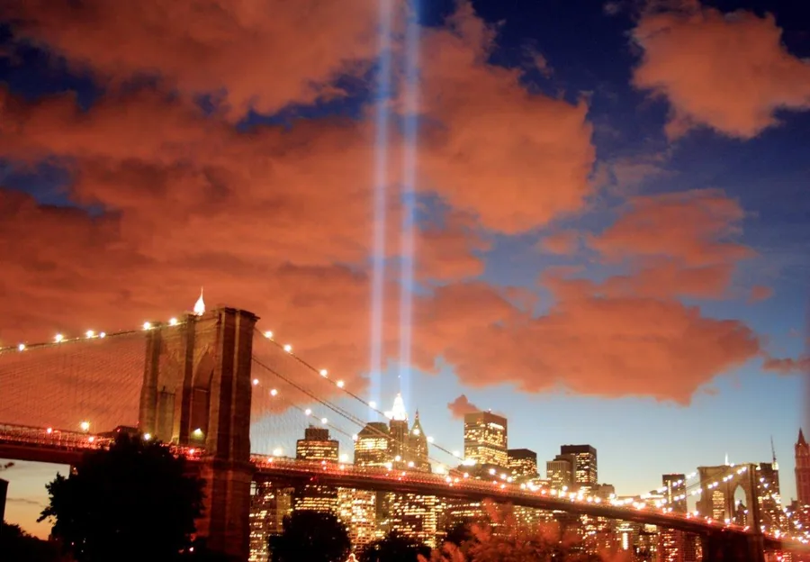 Brooklyn Bridge and Manhattan skyline at sunset with illuminated buildings and two light beams against orange and blue sky.