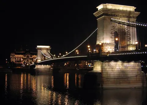 Nighttime view of an illuminated chain bridge with lit towers reflected in dark water, historic buildings visible in background.