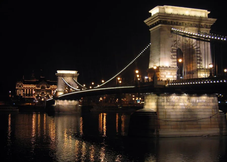 Nighttime view of an illuminated chain bridge with lit towers reflected in dark water, historic buildings visible in background.