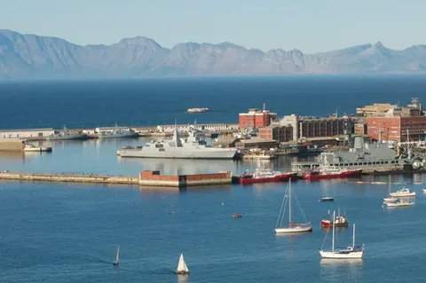 Scenic coastal harbor with white sailboats, red cargo ship, mountain range, and Mediterranean-style waterfront architecture.