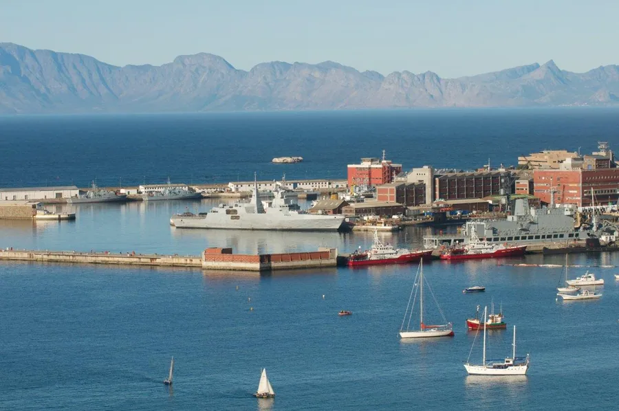 Scenic coastal harbor with white sailboats, red cargo ship, mountain range, and Mediterranean-style waterfront architecture.