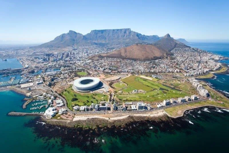 Aerial view of Cape Town showing Table Mountain, stadium, and coastal cityscape with turquoise ocean.