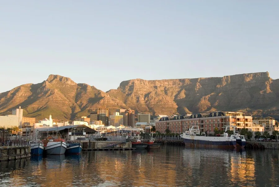 Cape Town waterfront with Table Mountain at sunset, boats docked in harbor, city buildings reflected in water.