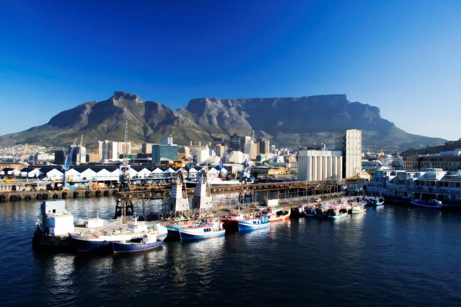 Aerial view of Cape Town harbour with Table Mountain, boats, and city skyline against blue sky.
