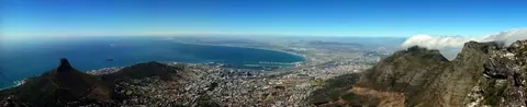 Panoramic aerial view of Cape Town with Table Mountain, surrounding peaks, blue ocean, and coastal cityscape under clear sky.