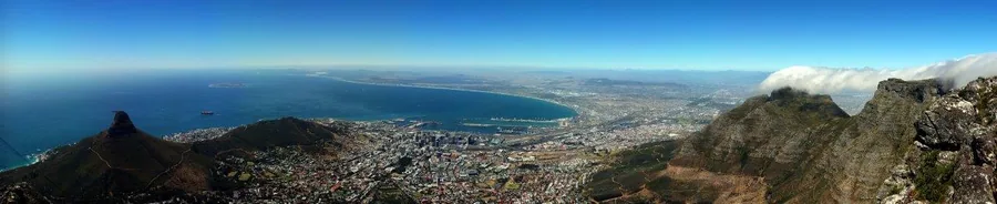 Panoramic aerial view of Cape Town with Table Mountain, surrounding peaks, blue ocean, and coastal cityscape under clear sky.