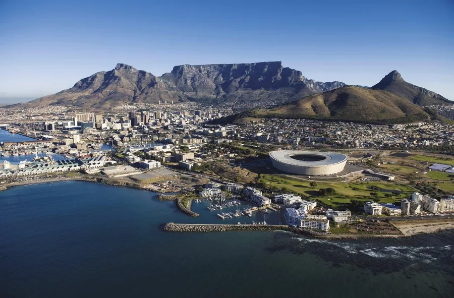 Aerial cityscape of Cape Town showing Table Mountain, stadium, harbor, and surrounding neighborhoods under clear blue sky.
