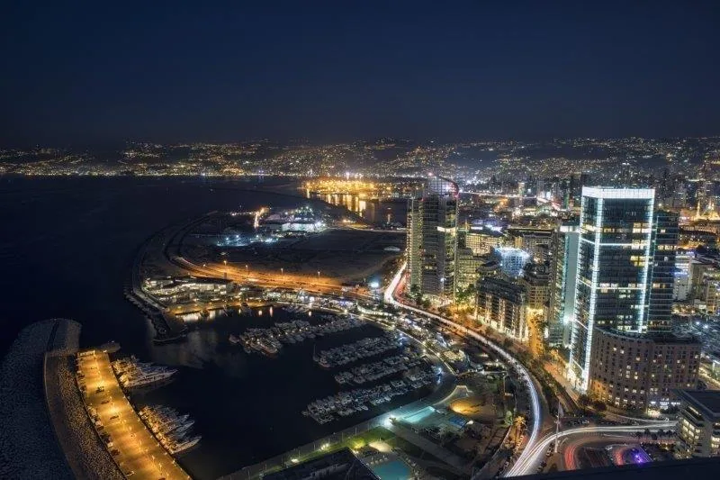 Nighttime aerial view of illuminated coastal city with harbor, buildings, and water reflections