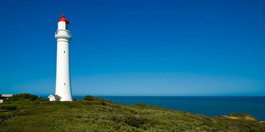 White lighthouse with red top on grassy clifftop overlooking blue ocean and clear sky.