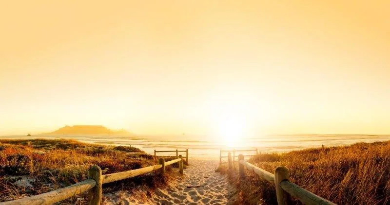 Wooden fence pathway on sandy beach at sunset with golden light and mountains in distance