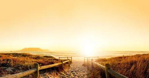Wooden boardwalk path leading to a golden sunrise over a beach with mountains in the distance.