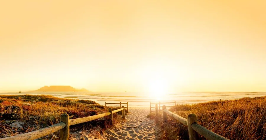 Wooden boardwalk path leading to a golden sunrise over a beach with mountains in the distance.