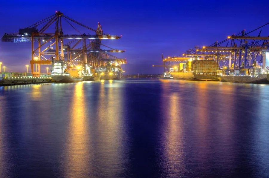 Nighttime shipping port with glowing yellow cranes reflected in blue water at dusk