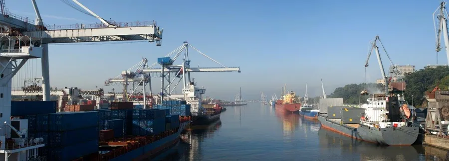 Busy harbour with blue cargo ships, industrial cranes, and calm water at sunrise in a coastal port.