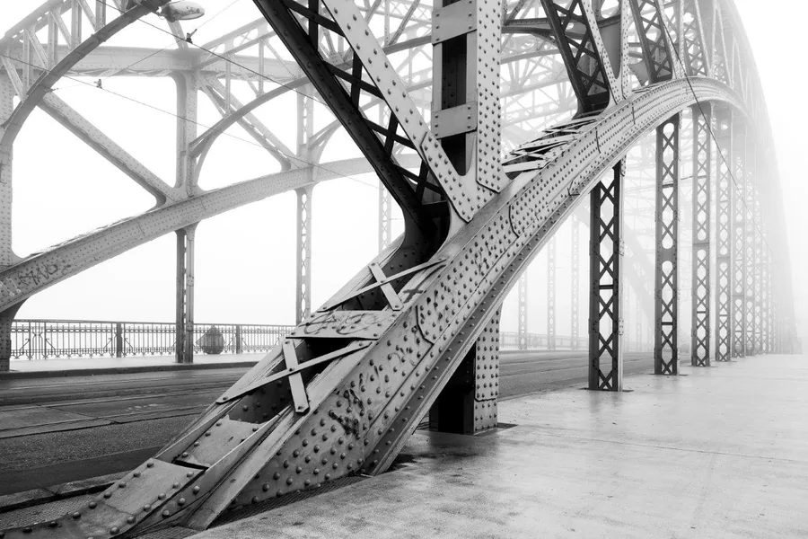 Black and white industrial bridge interior with steel beams, rivets, and geometric metal framework