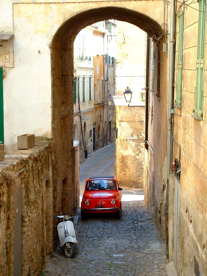Narrow Italian village street with arched stone passage, red vintage car, white scooter, and weathered ochre buildings.
