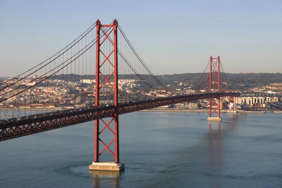 Red suspension bridge spanning blue water with Portuguese city skyline in background