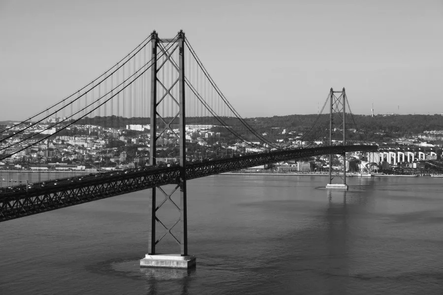Black and white image of a large suspension bridge over water with city buildings in the background
