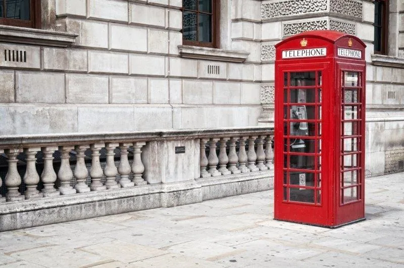 Red British telephone booth against cream stone building with ornate balustrade railing.