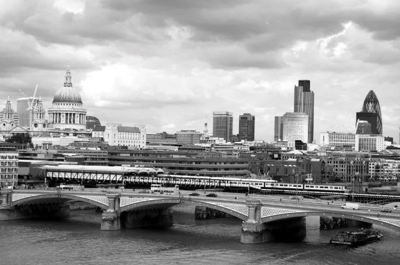 Black and white photograph of London skyline with St Paul's Cathedral, bridges over the Thames, and modern buildings under cloudy sky.