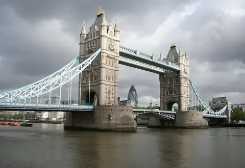 Tower Bridge London spanning the River Thames with ornate stone towers and light blue steel suspension cables against cloudy sky